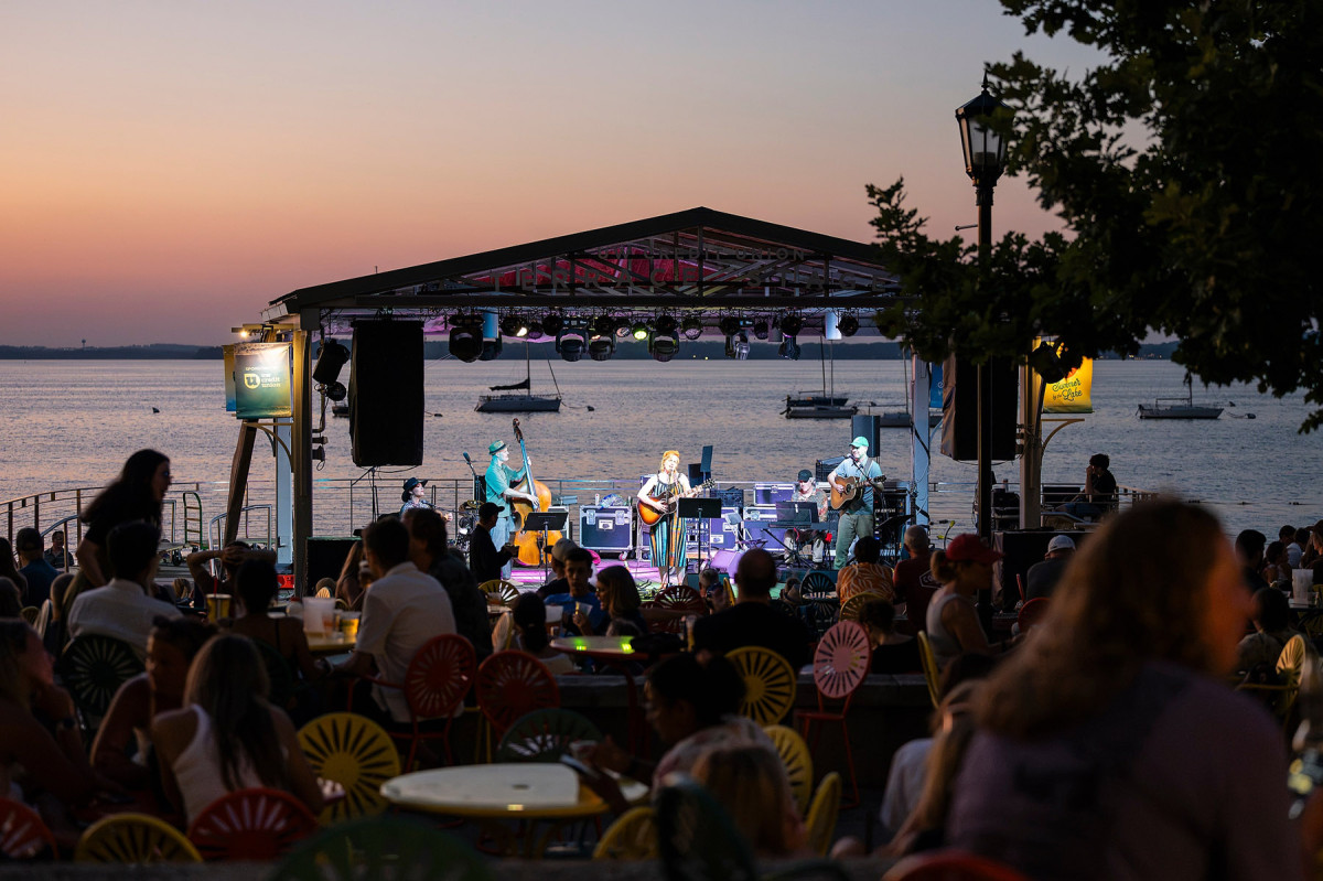 A band performs on the Terrace stage at night under colorful lights with a sunset Lake Mendota in the background