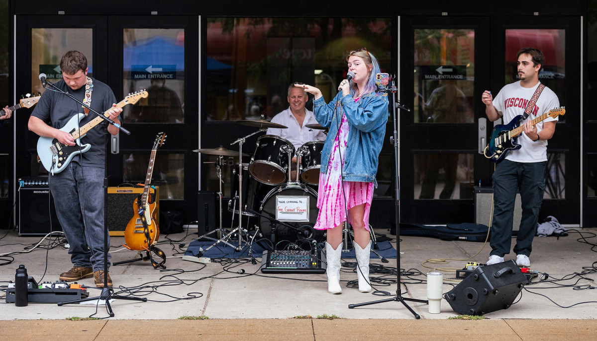 A four piece band, including a singer, two guitarists, and a drummer perform in front of Memorial Library