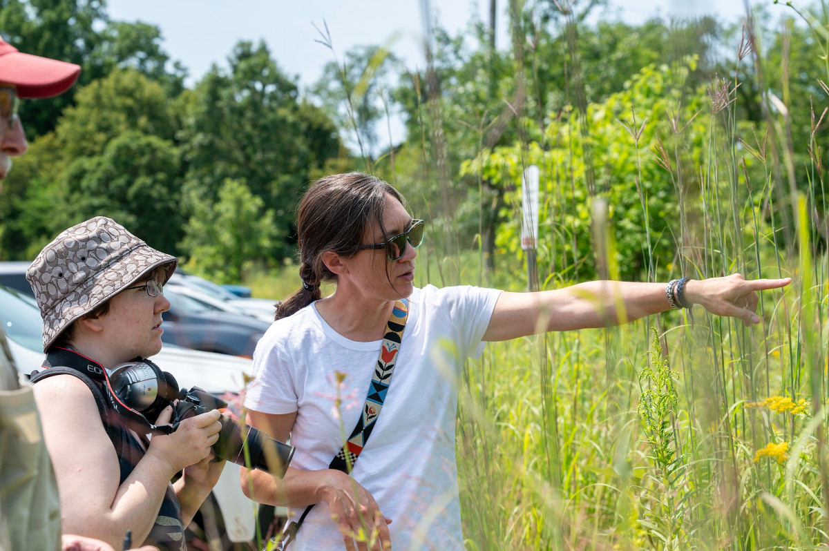 A woman points to a birds nest while another person stands by with a large camera ready to take a photo.