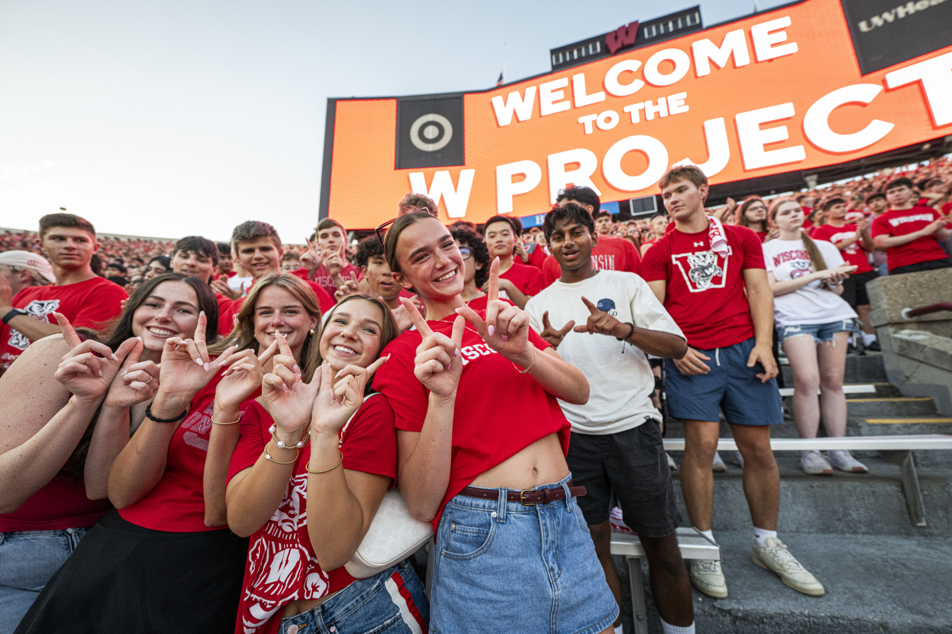 A group of UW students wearing Badger red hold up their hands in a W sign from the stands of a football stadium.