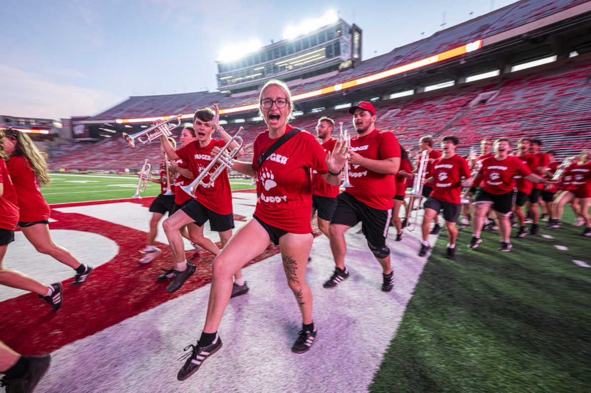 Members of the UW Marching band, all wearing smiles and Badger red tee shirts, march in formation holding their instruments in their arms.