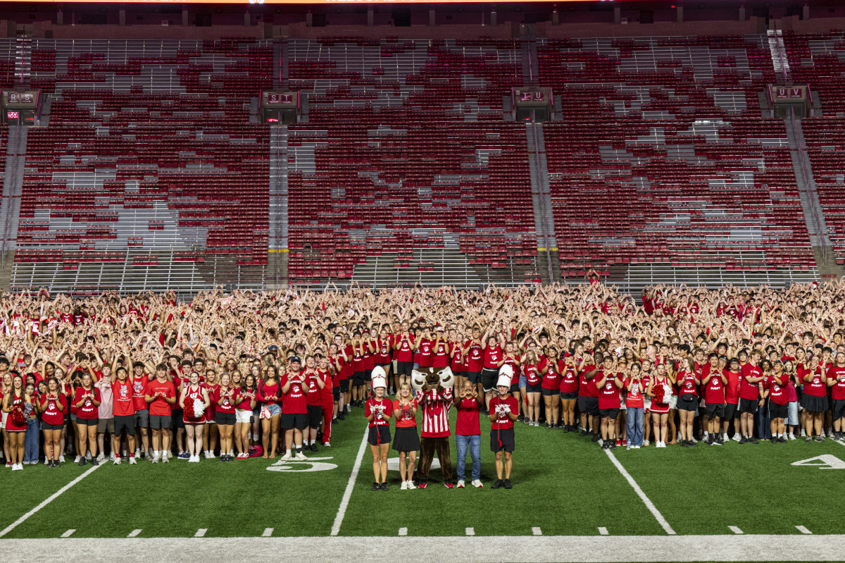 Mascot Bucky Badger poses with Wilcots, Ressor and two UW Marching Band drum majors in front of a large crowd of students.