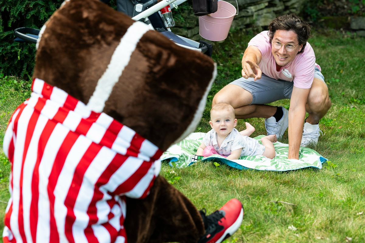 UW mascot Bucky Badger plays peek a boo with a baby who is laying on a blanket. The baby's father crouches nearby, pointing out Bucky.