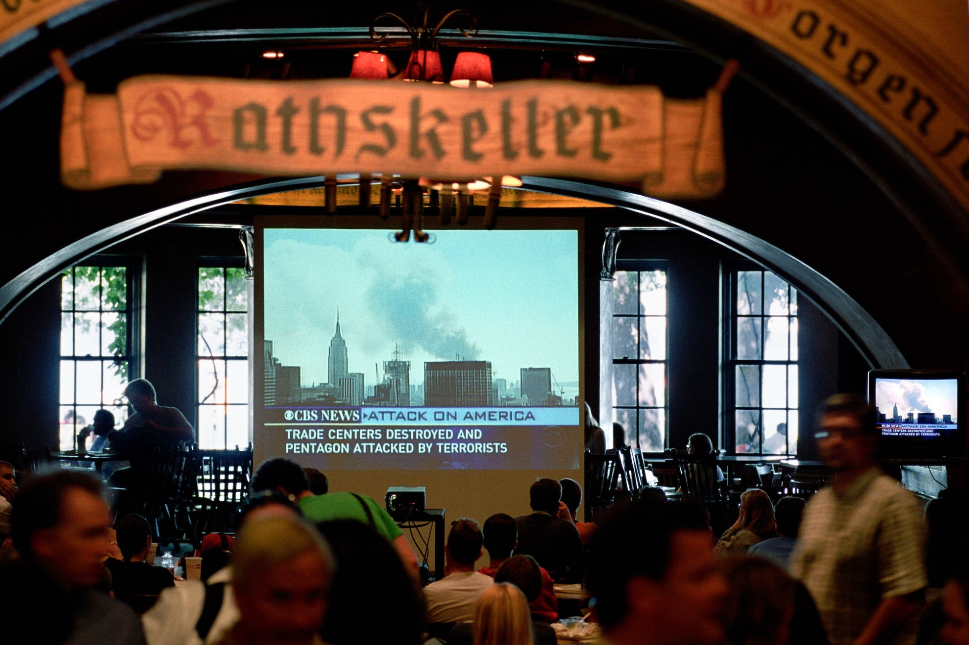 People sit and watch a projector screen in the Rathskeller. The screen shows a skyline with smoke in the air, with the text "Attack on America."
