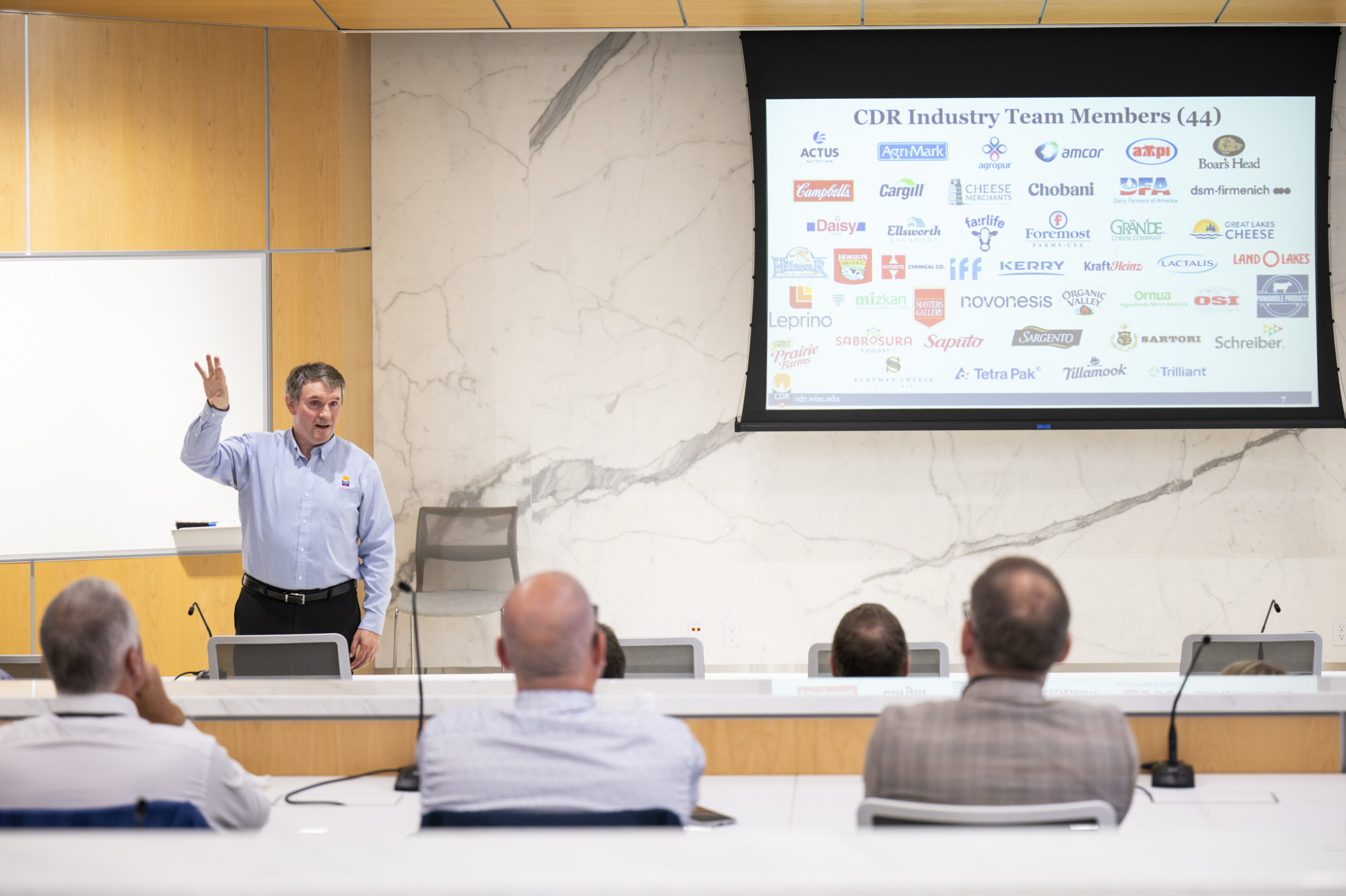 Dairy research center director John Lucey presents to a crowd in front of a screen that displays corporate sponsor logos. 