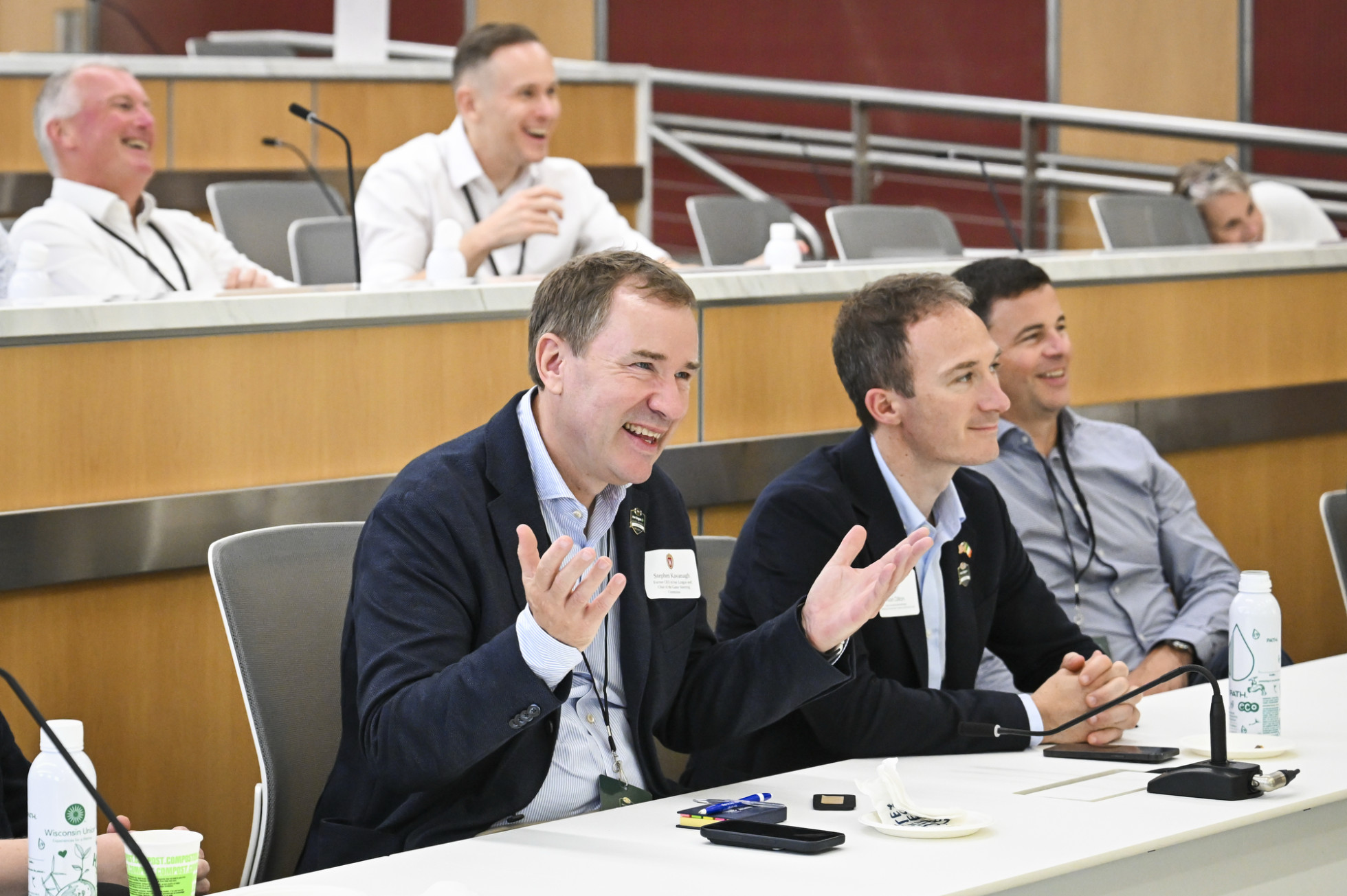 Stephen Kavanagh, smiling widely and sitting at a table, raises his hands up while asking a question. 