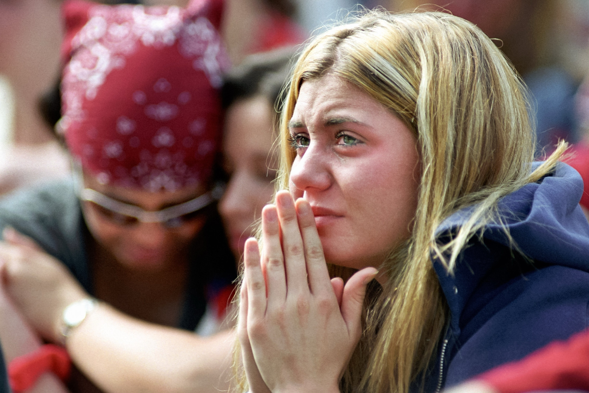 A student, with teary eyes, holds her hands together, with people embracing behind her, during a moment of silence.