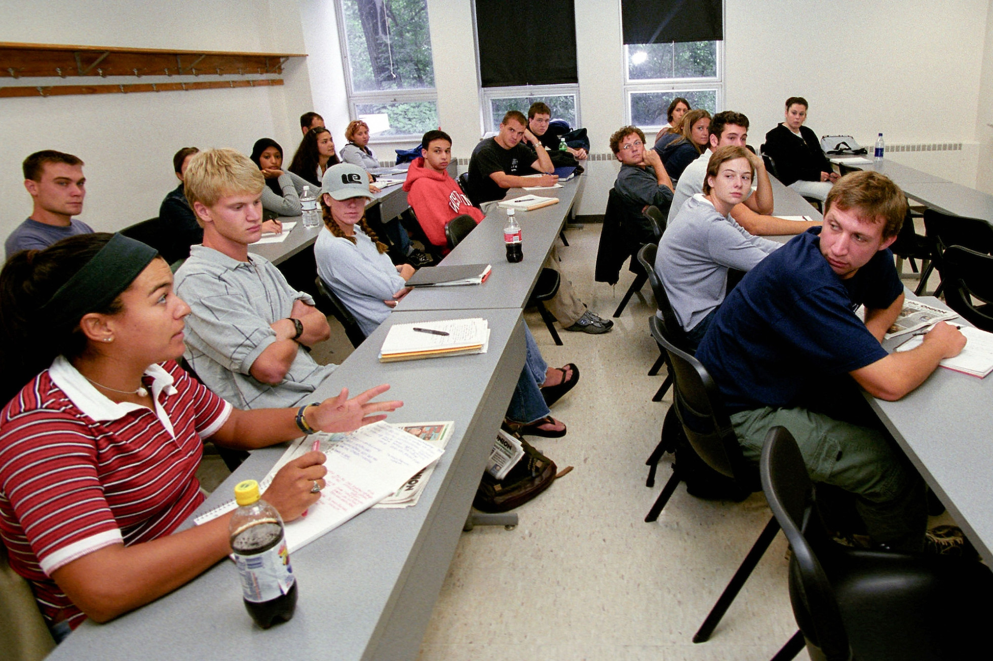 Students sitting at rows of tables in a classroom speak to each other.
