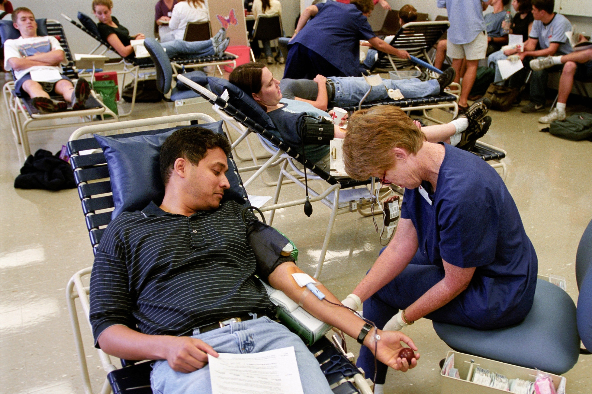 A medical professional attends to equipment as a man is donating blood, with others doing the same in the background.