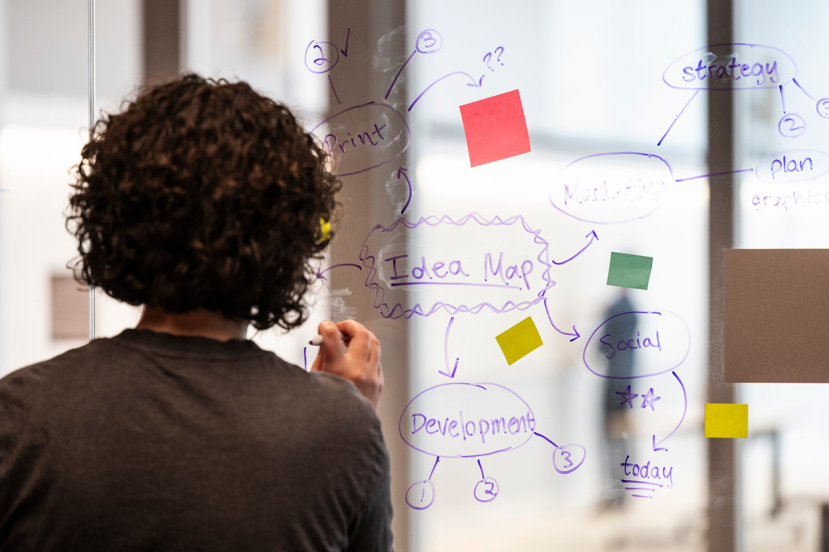 A women stands facing a see through white board. She makes notes on the board and adds colorful post it notes to the board.