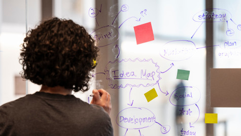 A women stands facing a see through white board. She makes notes on the board and adds colorful post it notes to the board.