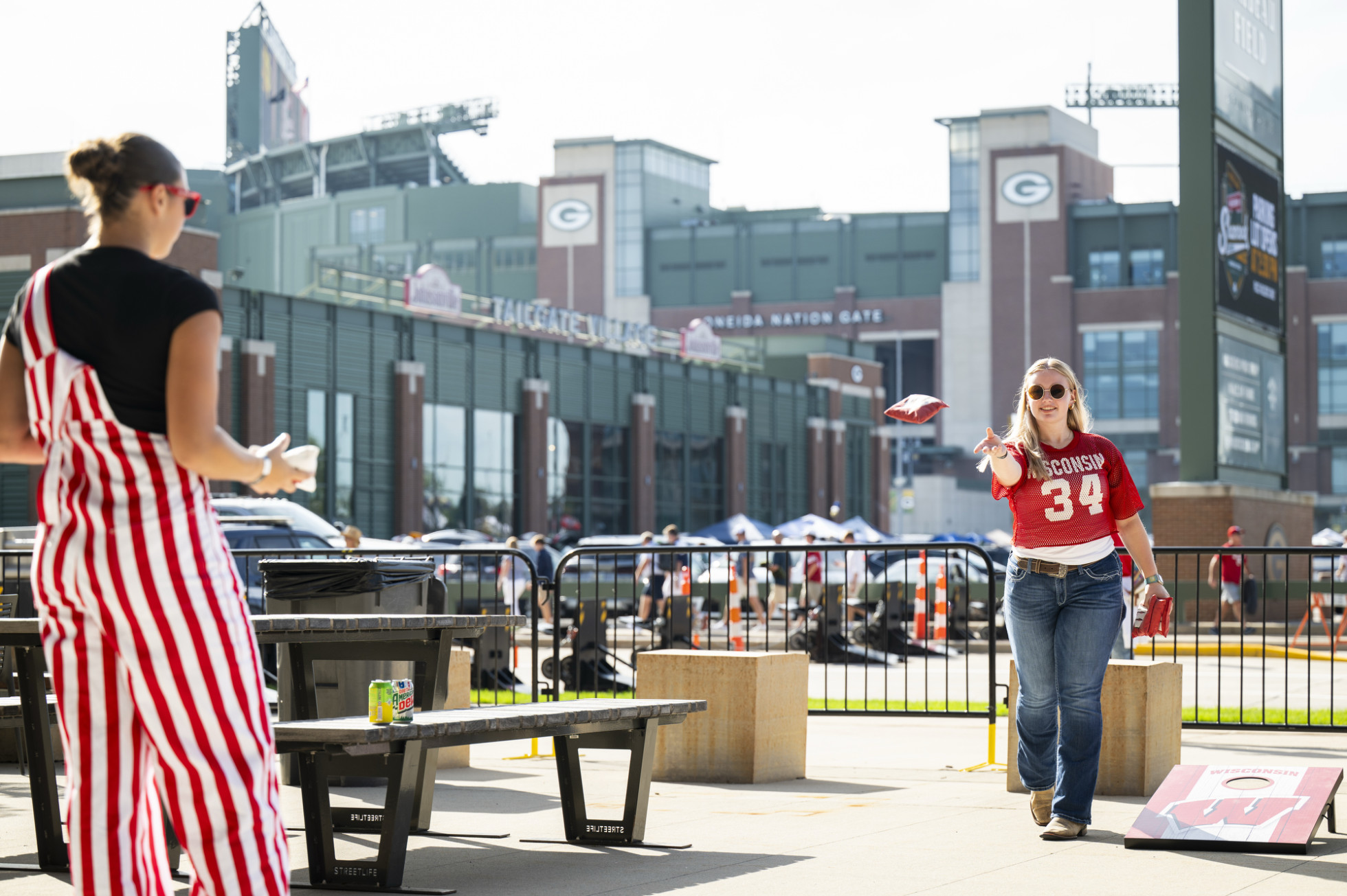 Two tailgaters wearing Badger gear play bean bag toss, with Lambeau Field in the background.
