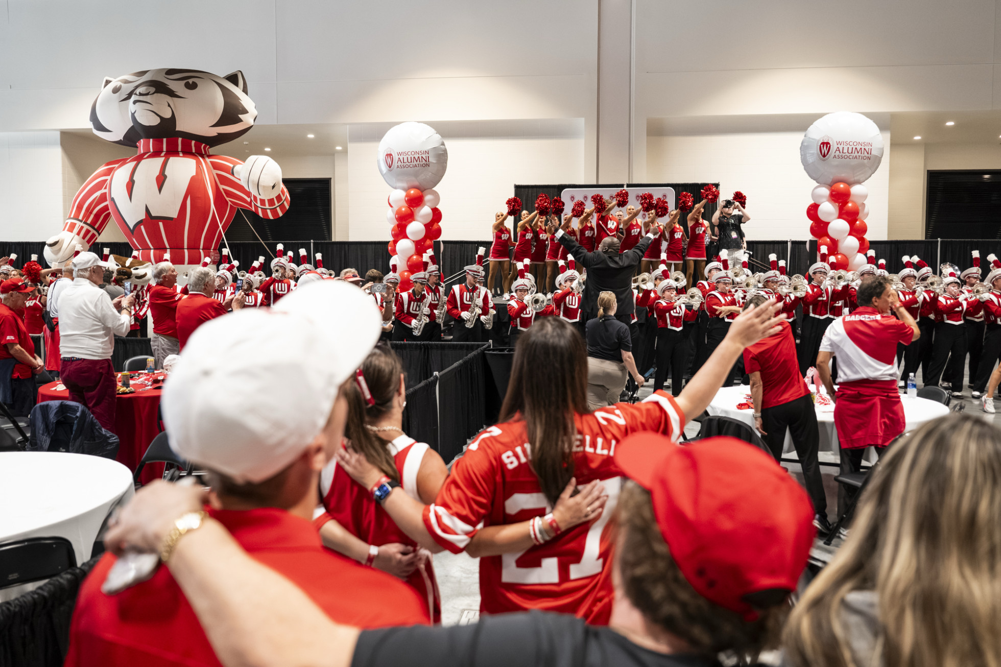 Badger fans embrace each other during the singing of "Varsity" in an auditorium, with the UW Marching Band and Spirit Squad on stage. 