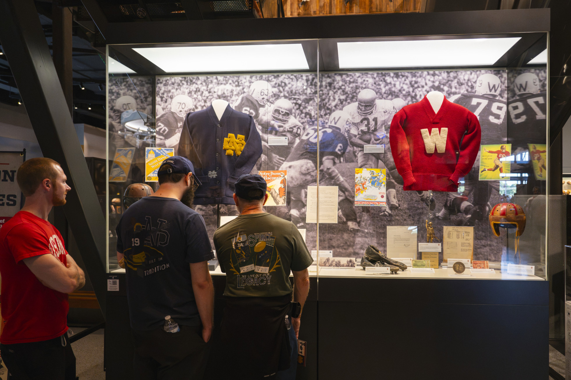 Three people look at a glass display that shows early football uniforms and other artifacts from Wisconsin and Notre Dame.
