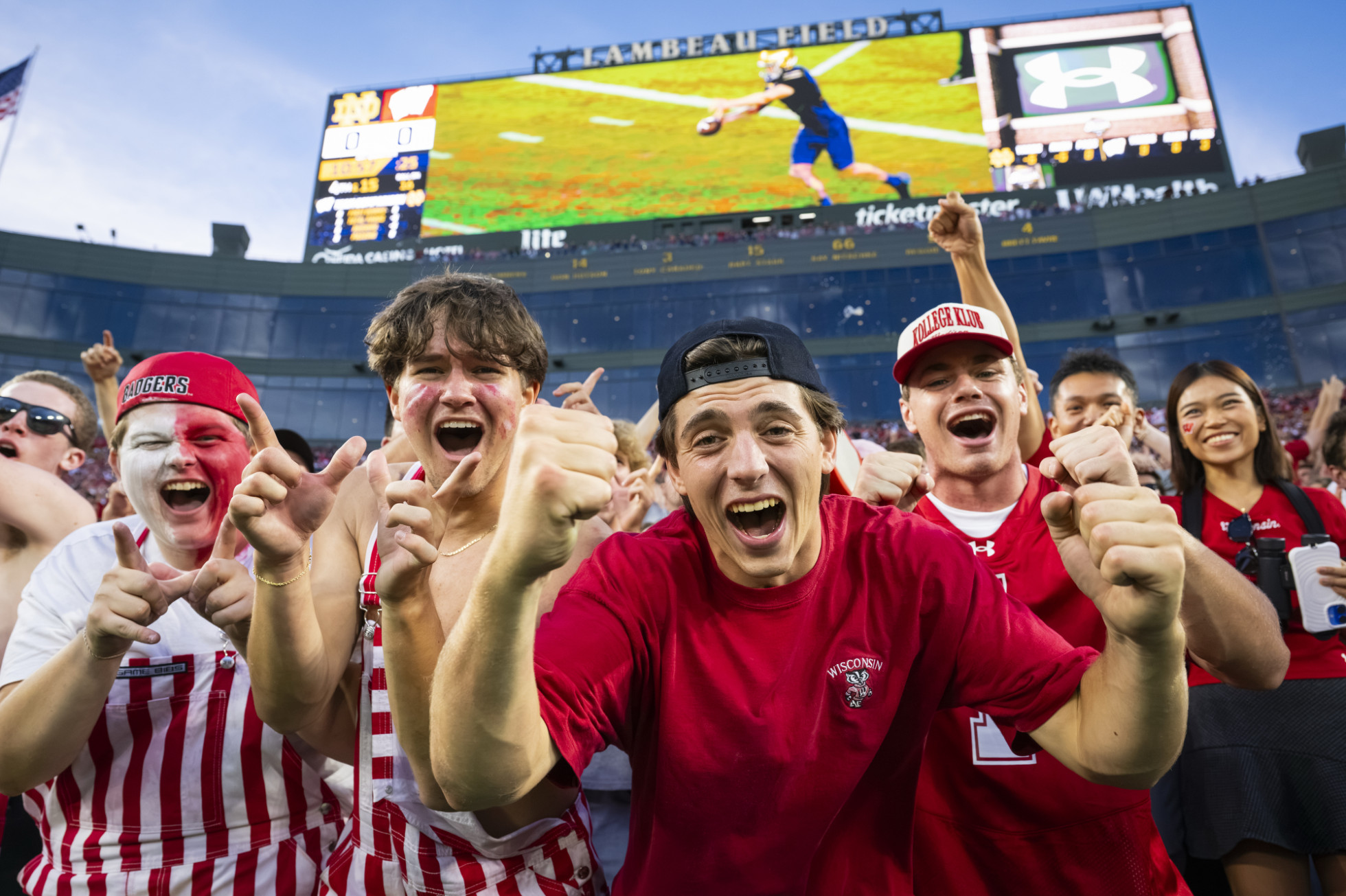 Football fans wearing Badger gear cheer at the camera, with the Lambeau Field scoreboard in the background. 