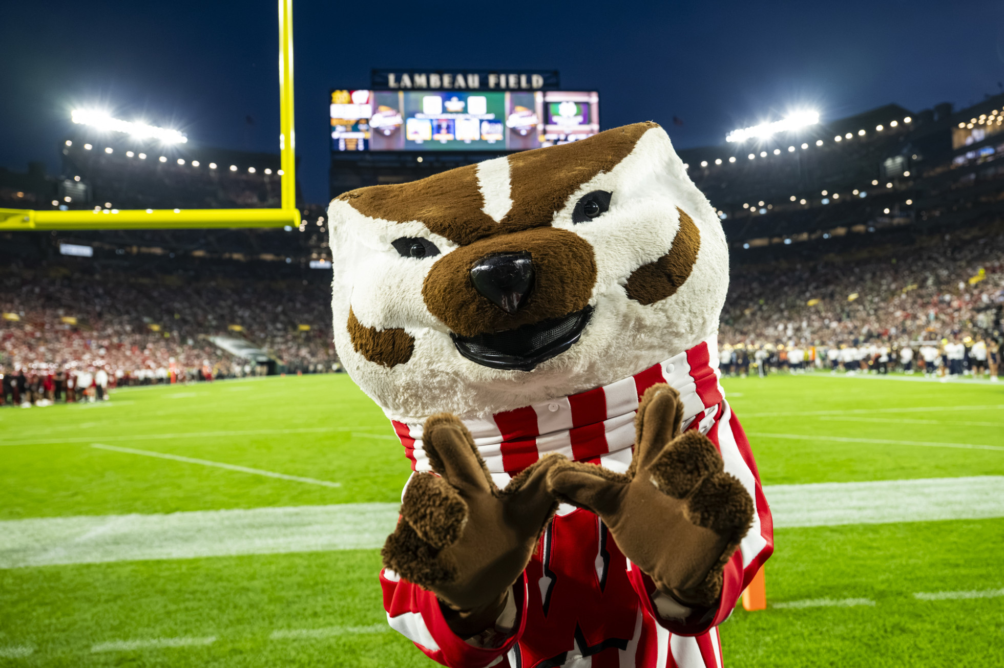 Bucky Badger flashes a W with his hands in front of a football field. 