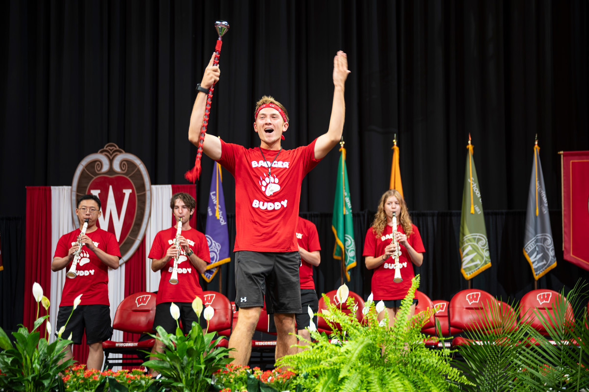 A drum major in a red "Badger Buddy" T-shirt smiles and raises his arms on stage, in front of several clarinet players.