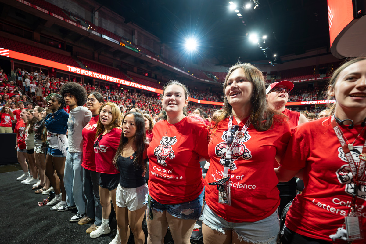 A row of students wearing Badger gear wrap their arms around each other’s backs.