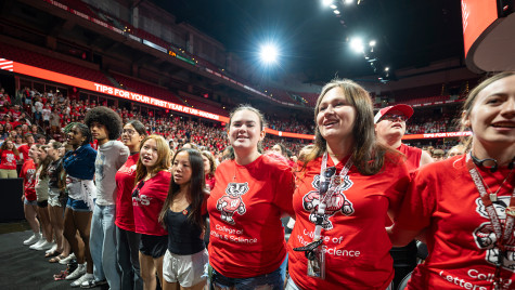 A row of students wearing Badger gear wrap their arms around each other’s backs.