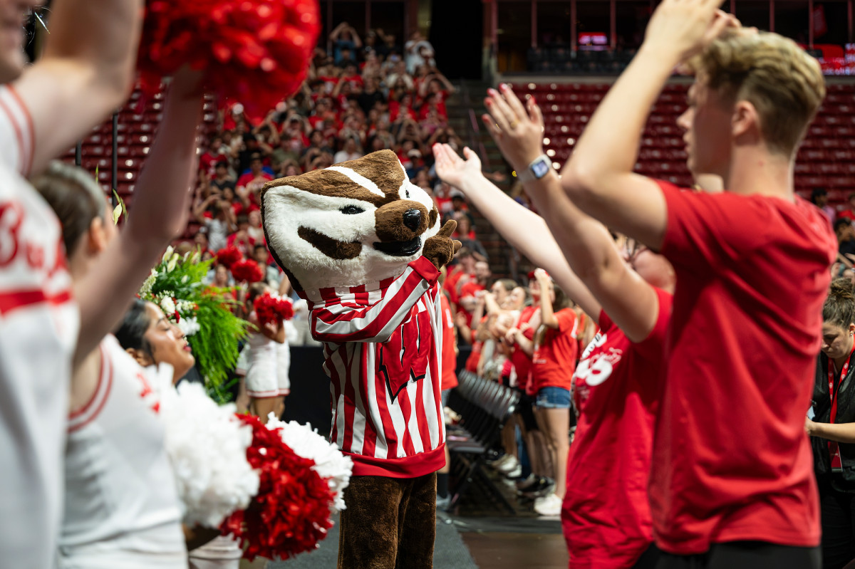 The Bucky Badger mascot walks down an arena aisle, wearing a red-and-white striped sweater and greeted by students and cheerleaders.
