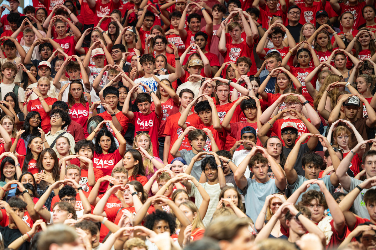 A large crowd of students dressed in red make a motion over their heads to the “By the light of the moon” song.