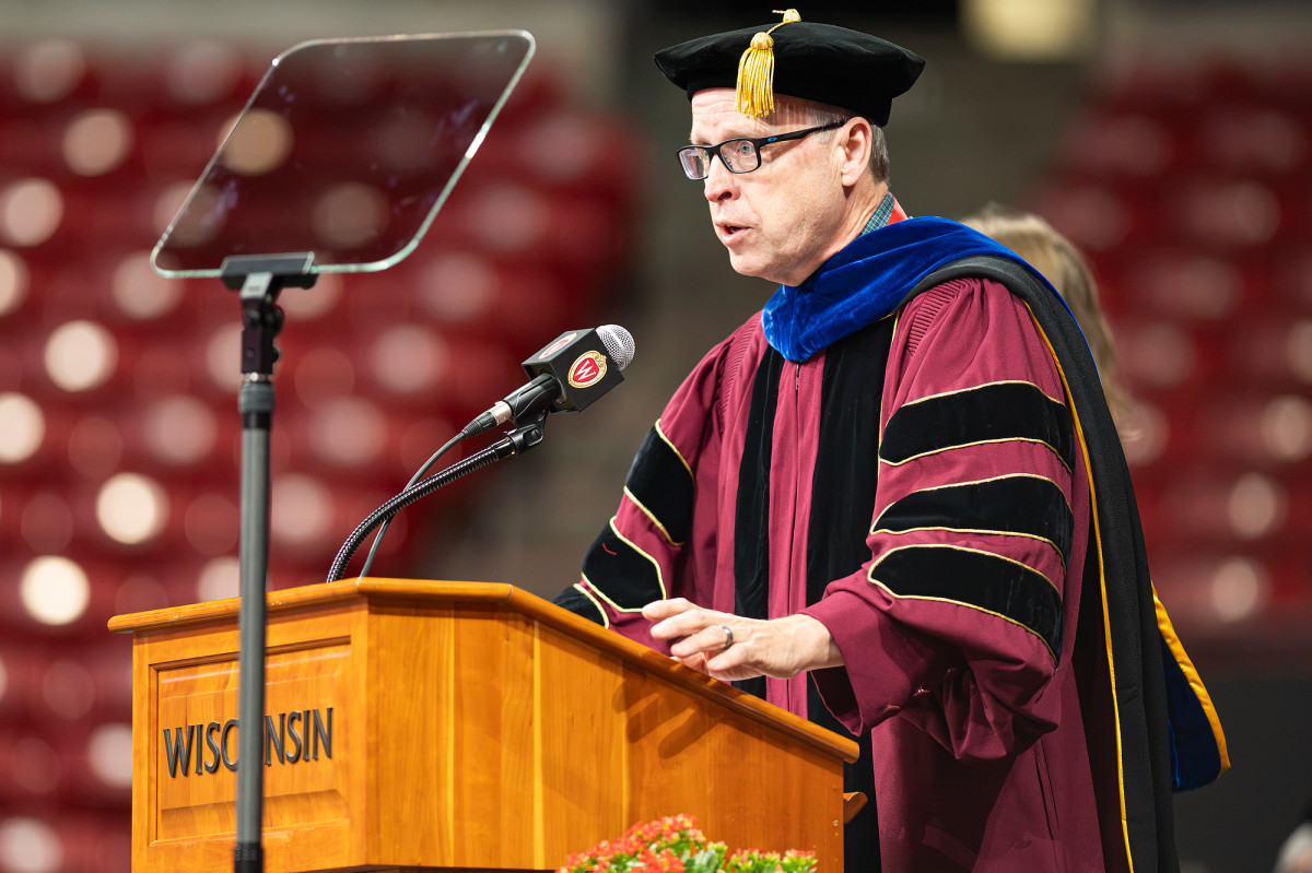 Provost John Zumbrunnen, in academic regalia, addresses the crowd from behind a wooden lectern emblazoned with the word “Wisconsin.”