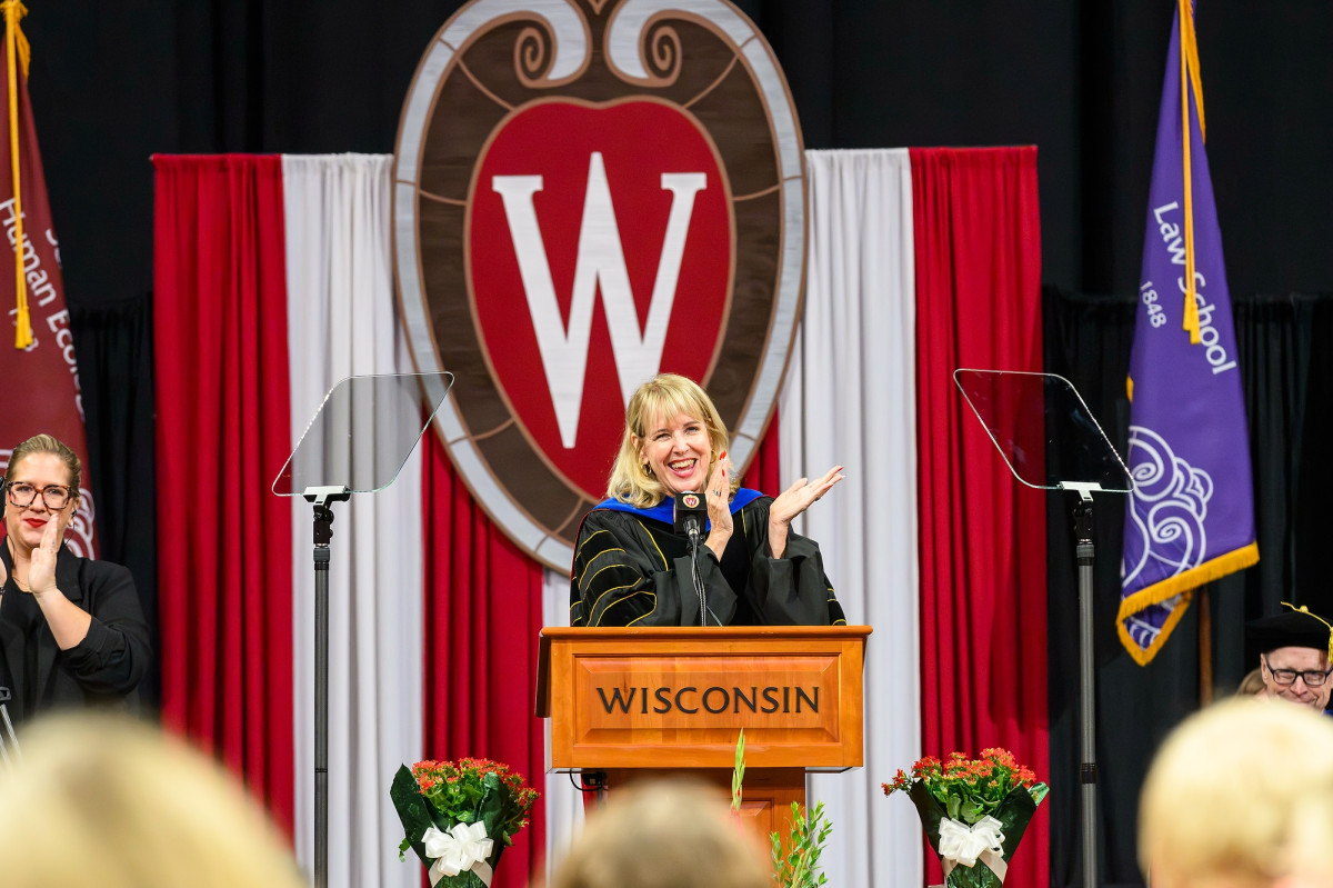 Vice Chancellor Lori Reesor, in academic regalia, smiles and claps from behind a wooden podium.