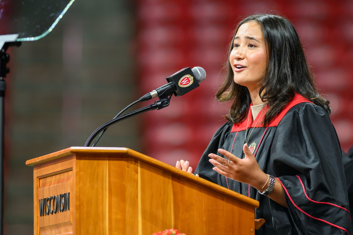 Student speaker Azi Fanale, in academic regalia, addresses the crowd from behind a wooden podium.