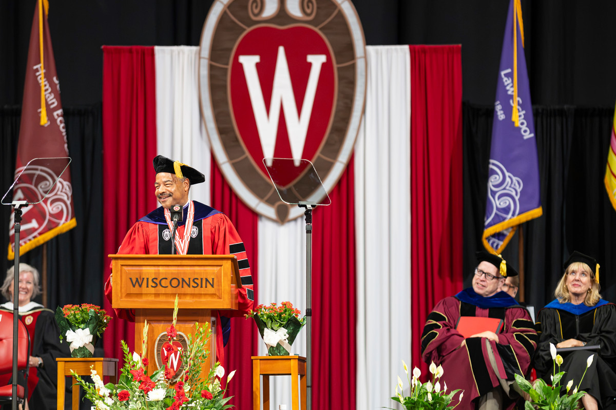 Interim Chancellor Eric Wilcots, in academic regalia, addresses the crowd from behind a wooden lectern emblazoned with the word “Wisconsin.”