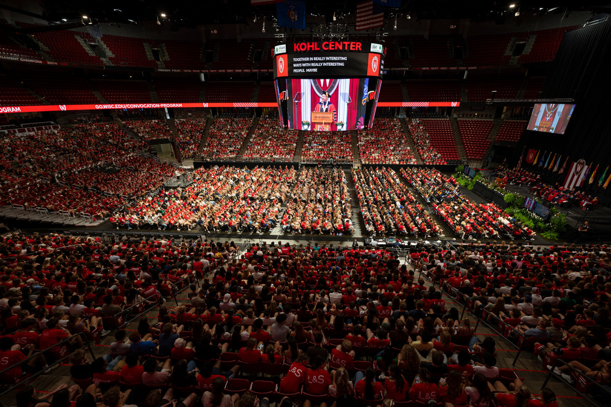 A high-angle view of the Kohl Center packed with new students, with a scoreboard displaying a live feed of the speaker at the podium.