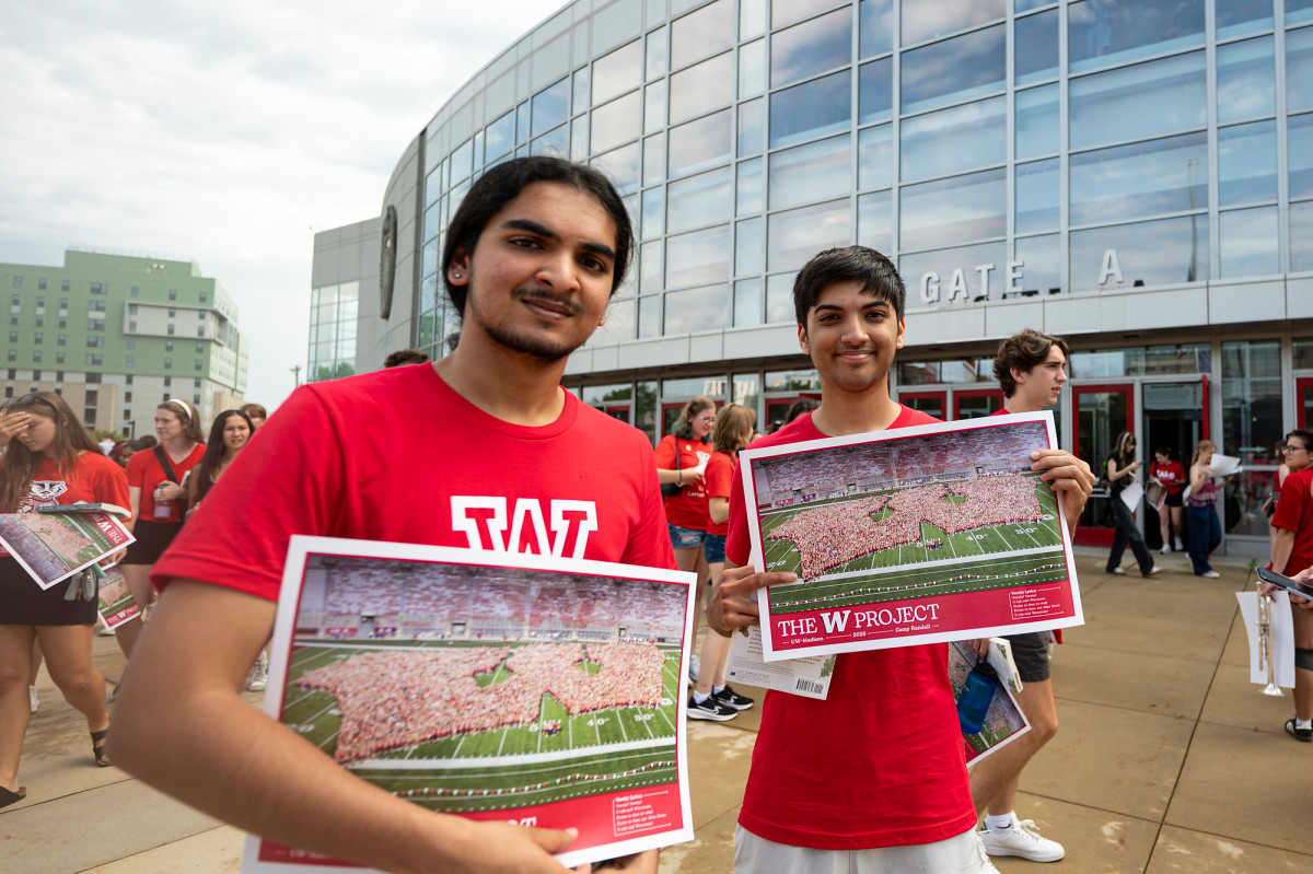 Outside of the Kohl Center, two students hold up posters with a photo of students gathered on the Camp Randall Field in the shape of a W.