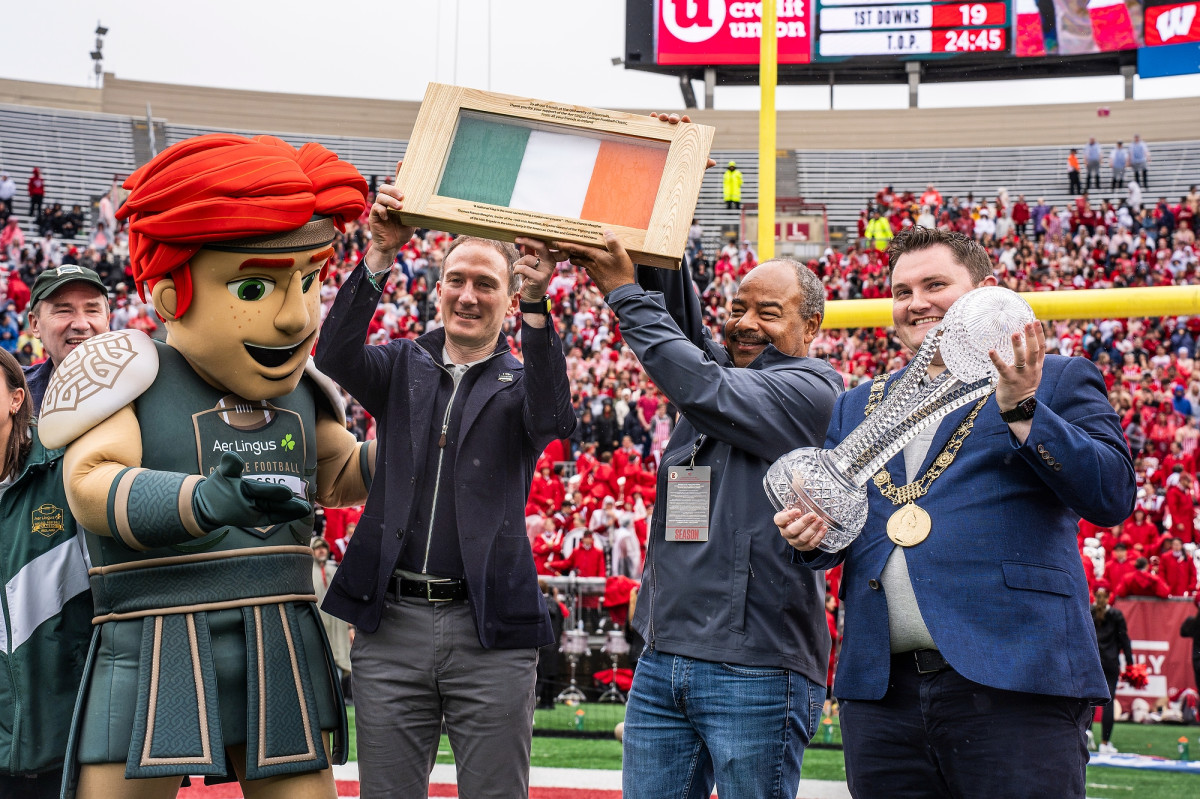 The Celtic the Brave mascot, Minister Alan Dillon, interim Chancellor Eric Wilcots and Lord Mayor Daryl Barron hold up the Irish flag and a trophy on the Camp Randall field.