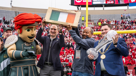 The Celtic the Brave mascot, Minister Alan Dillon, interim Chancellor Eric Wilcots and Lord Mayor Daryl Barron hold up the Irish flag and a trophy on the Camp Randall field.