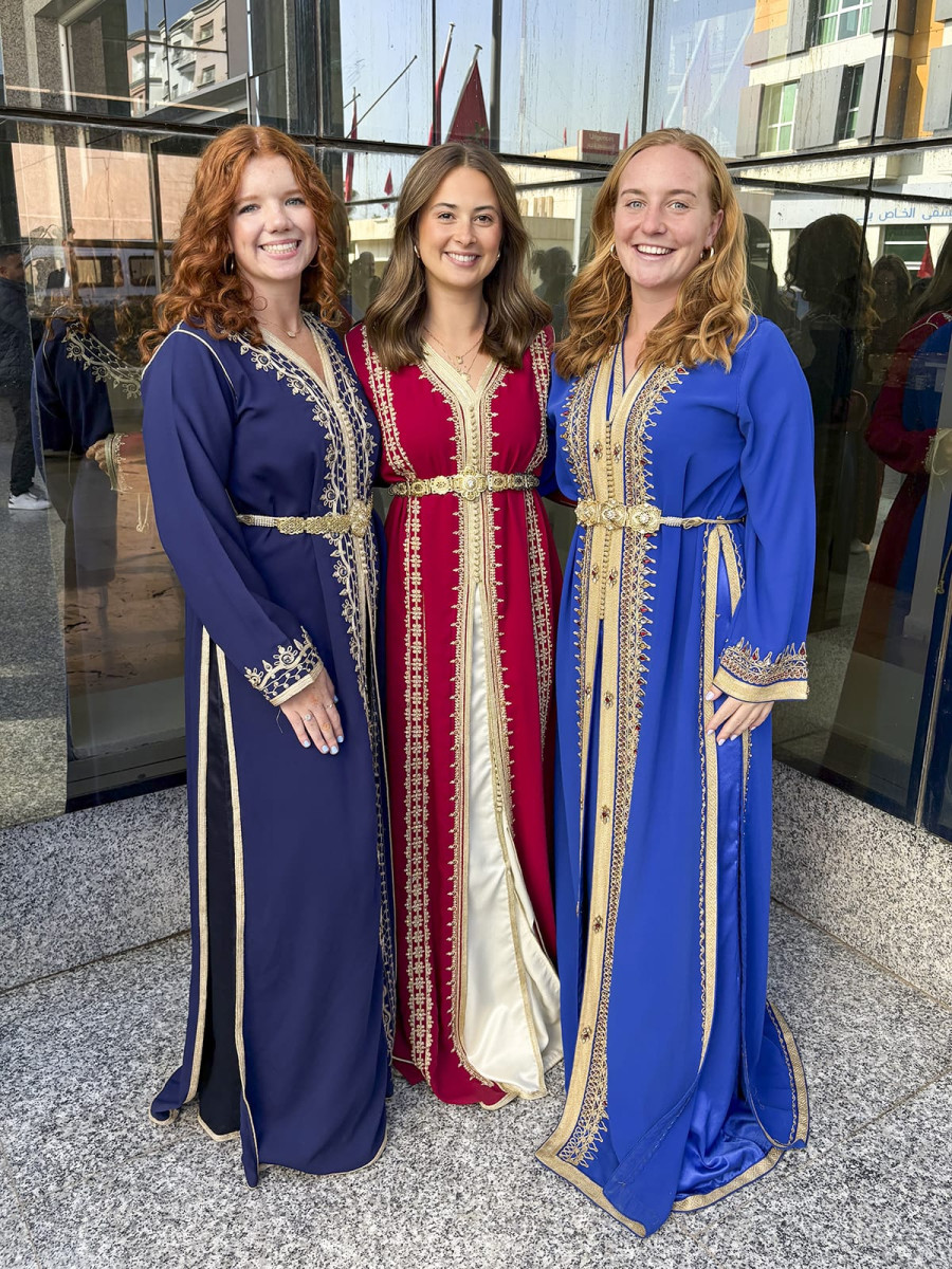 Three smiling women stand side-by-side wearing traditional Moroccan kaftans with ornate gold belts and embroidery in dark blue, maroon, and royal blue.