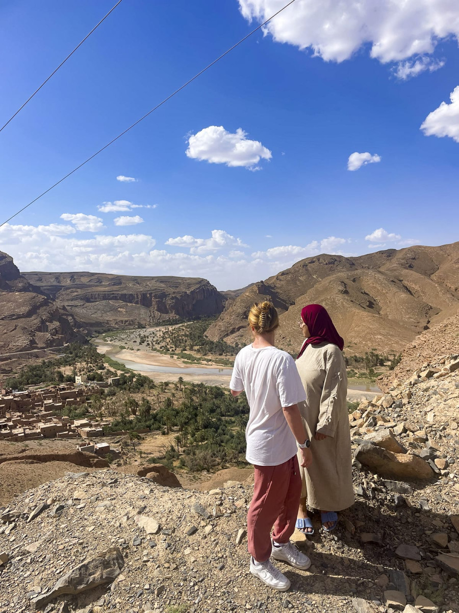Two women stand on a rocky overlook facing a desert river valley with sparse palm trees and surrounded by mountains under a bright blue sky.