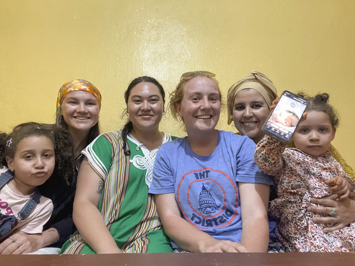 Four women and two young children sit together indoors against a yellow wall, with one child holding up a smartphone displaying a photo.