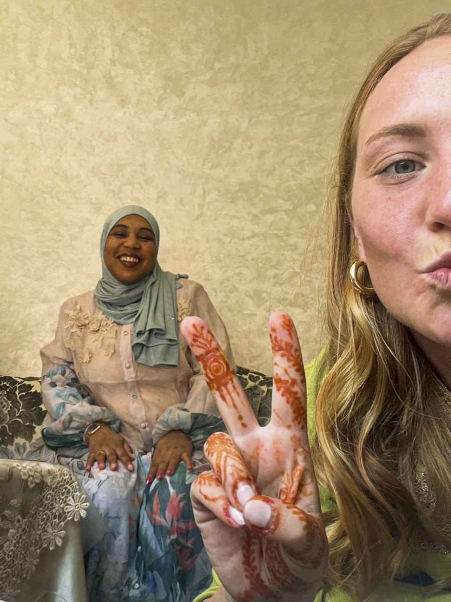 A close-up selfie of a woman displaying detailed henna designs on her hand in a peace sign gesture, with a smiling woman wearing a hijab sitting in the background.