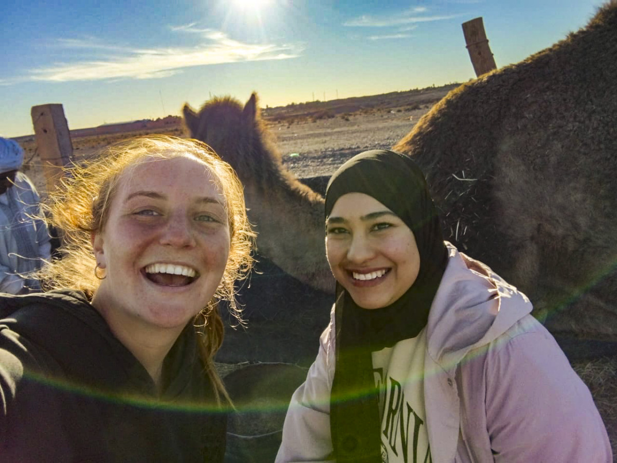 Two smiling women take an outdoor selfie in a sunny desert setting with camels standing behind them.