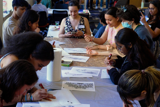 A group of students sit around a table and decorate each others hands with henna designs