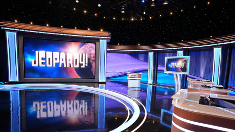 The empty studio set of the Jeopardy! game show features a large illuminated screen displaying the logo, a central host lectern, and contestant podiums under blue and purple studio lighting.