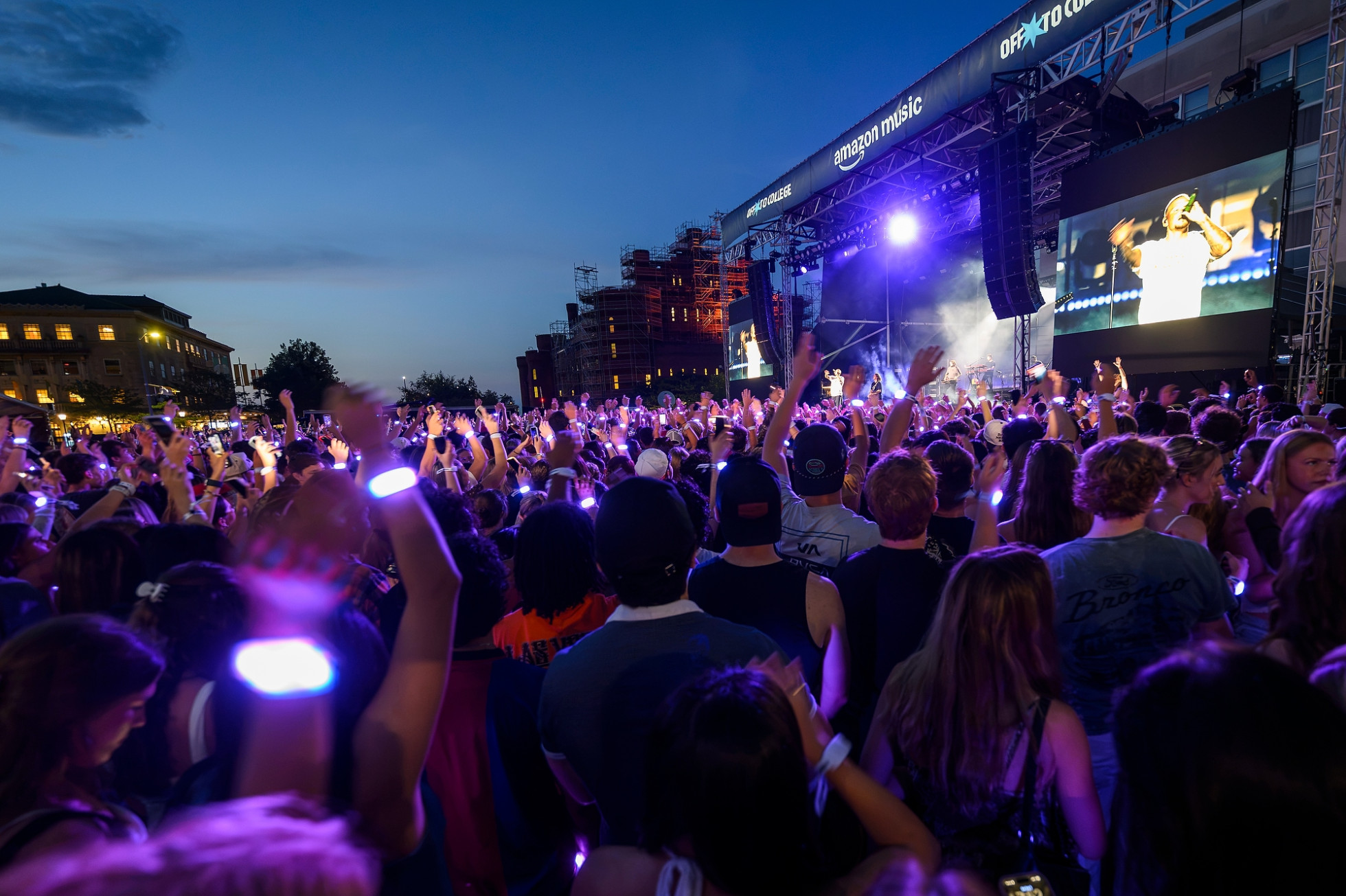 Concert crowd members, shown from behind, wave their arms in front of the stage, with the Red Gym and Memorial Union in the background.