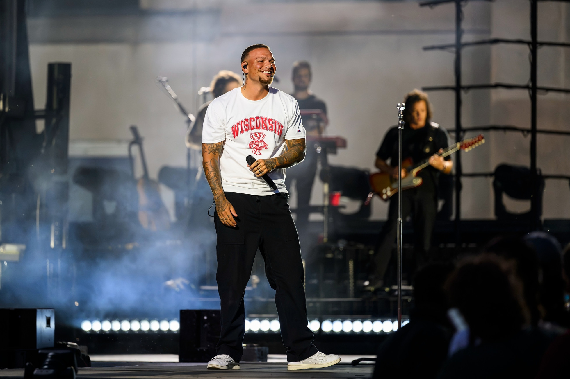 Kane Brown, wearing a Badger T-shirt, smiles and holds a microphone on stage.