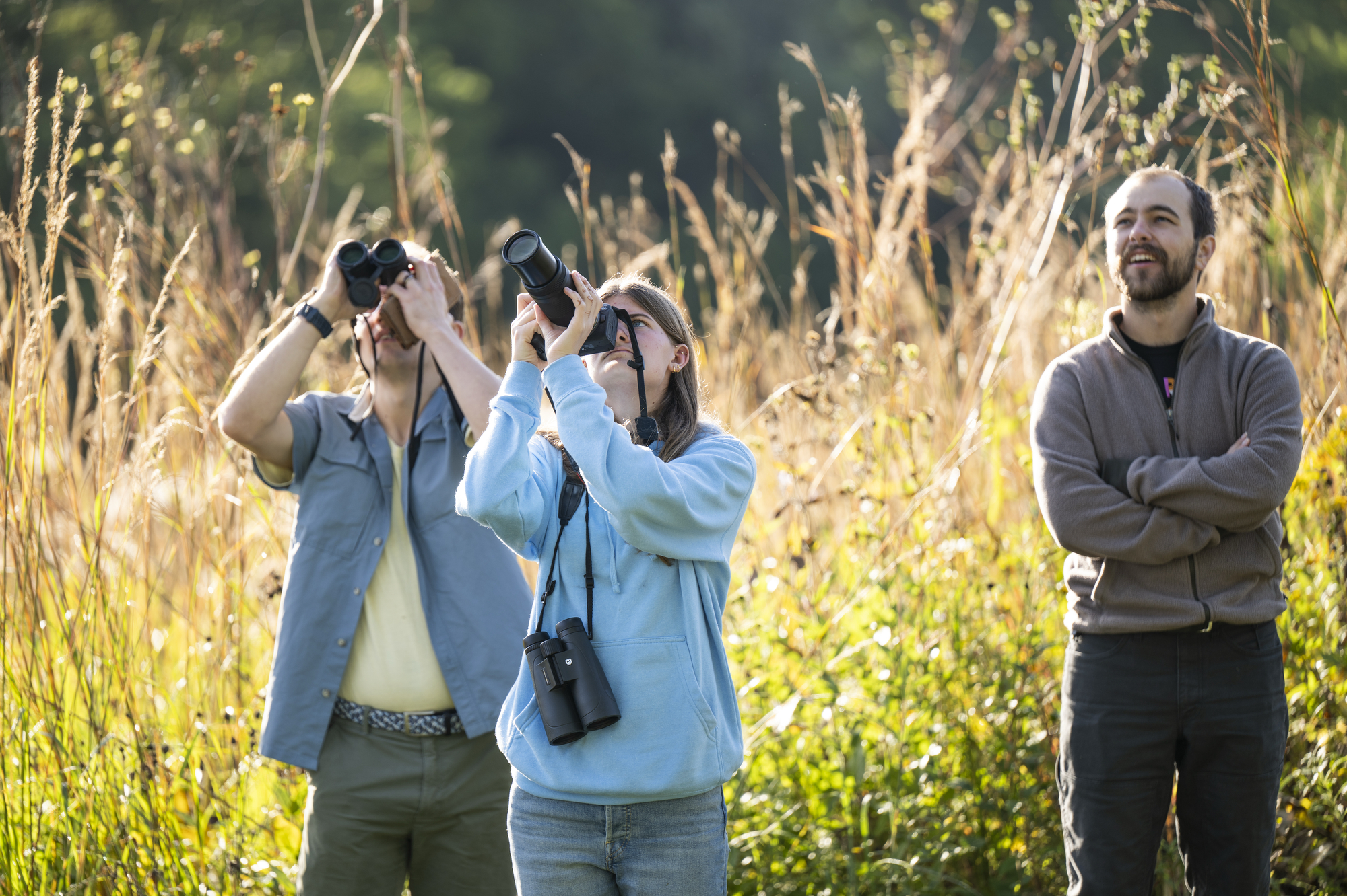 A group of people stand by a native prairie field and look through binoculars 
