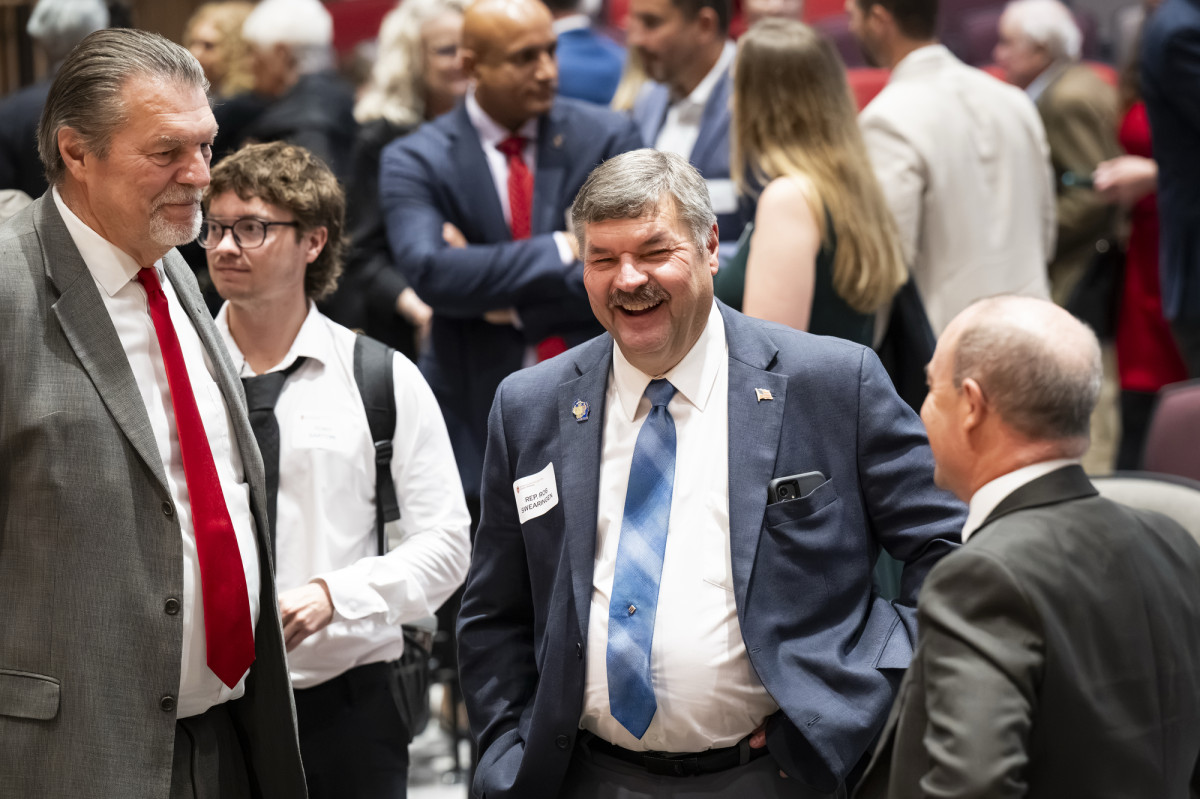 Three men wearing suits talk together in a group of many people gathered.