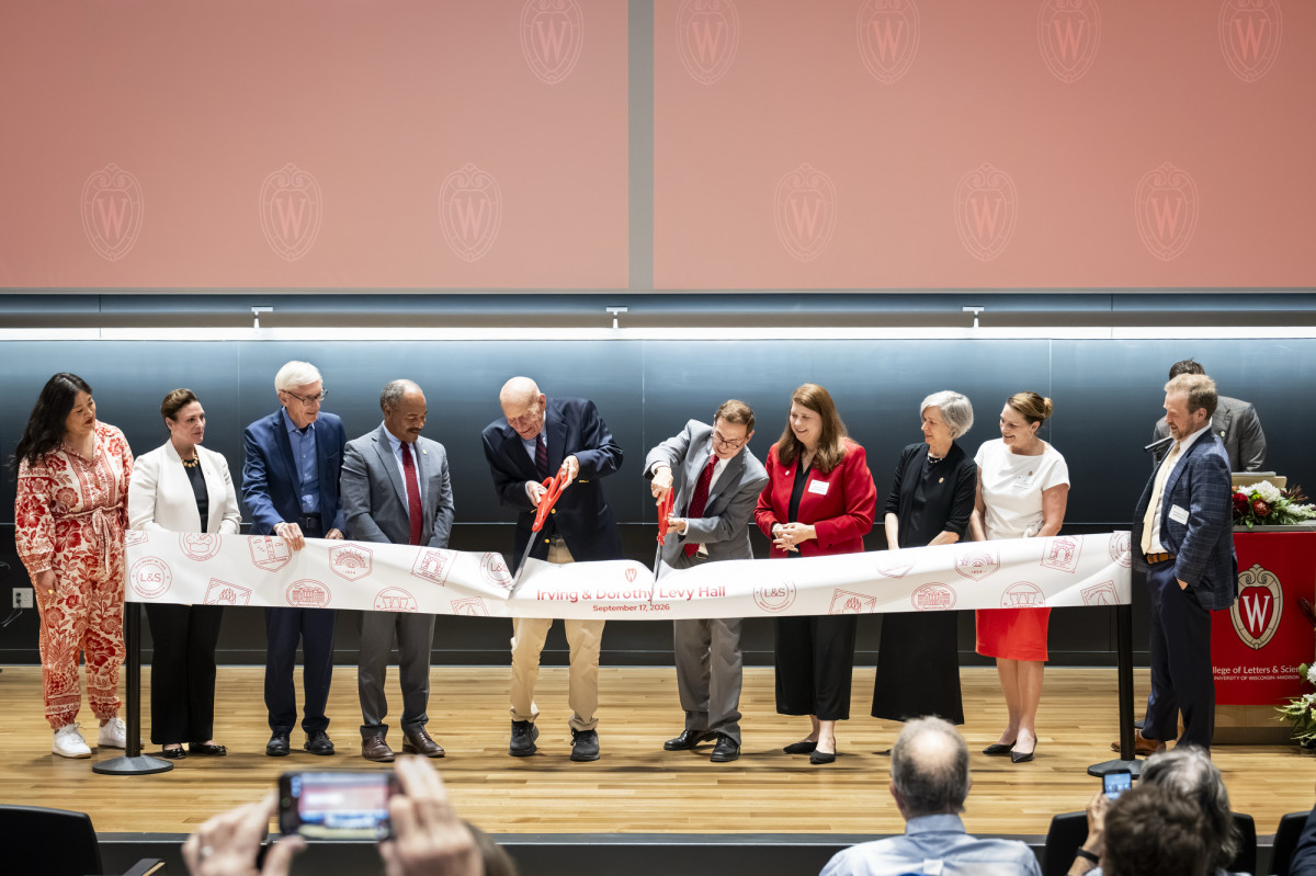 Two men with large ceremonial scissors cut a banner on stage at a ribbon-cutting ceremony as other dignitaries look on.