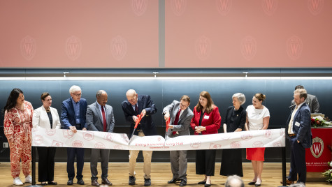 Two men with ceremonial scissors cut a large banner on a stage while other dignitaries look on.