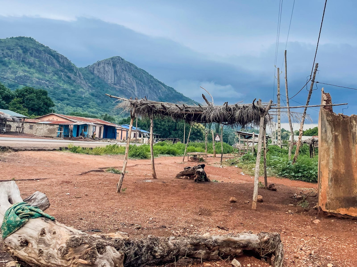 A thatched wooden shelter stands on reddish dirt in a rural village, set against green mountains under an overcast sky.