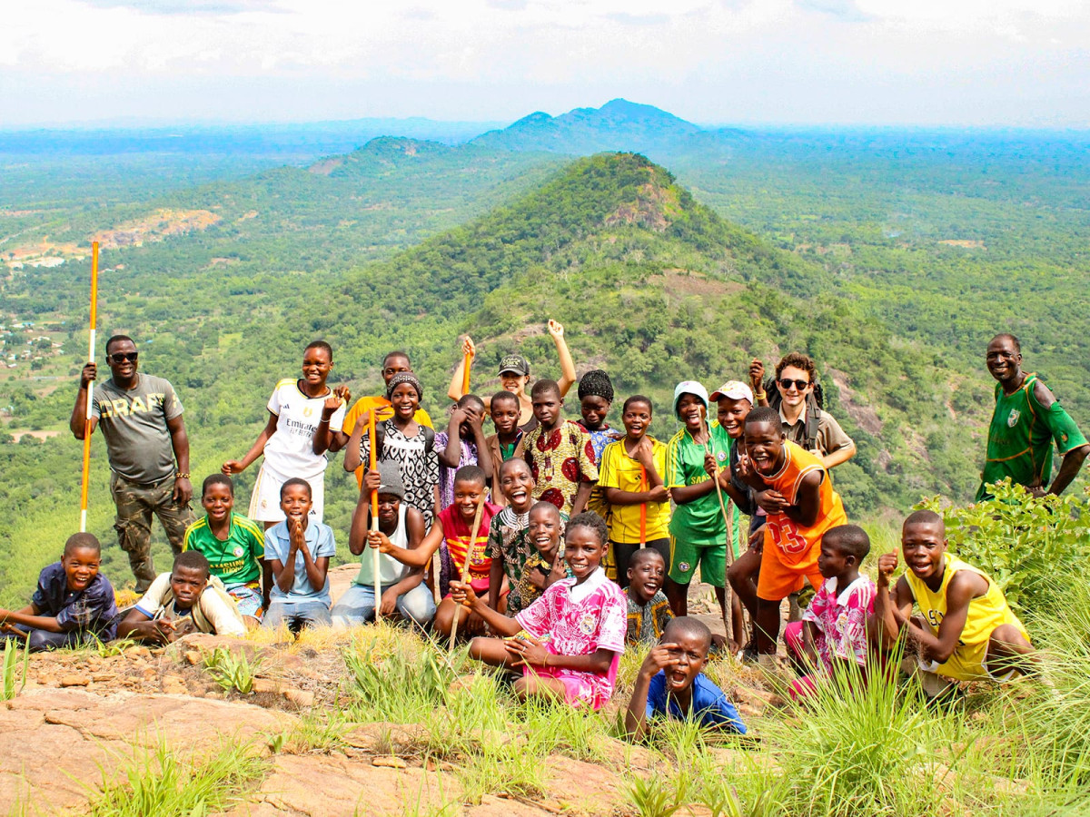 A large group of smiling children and adults pose together on a rocky hill summit overlooking an expanse of lush green countryside.