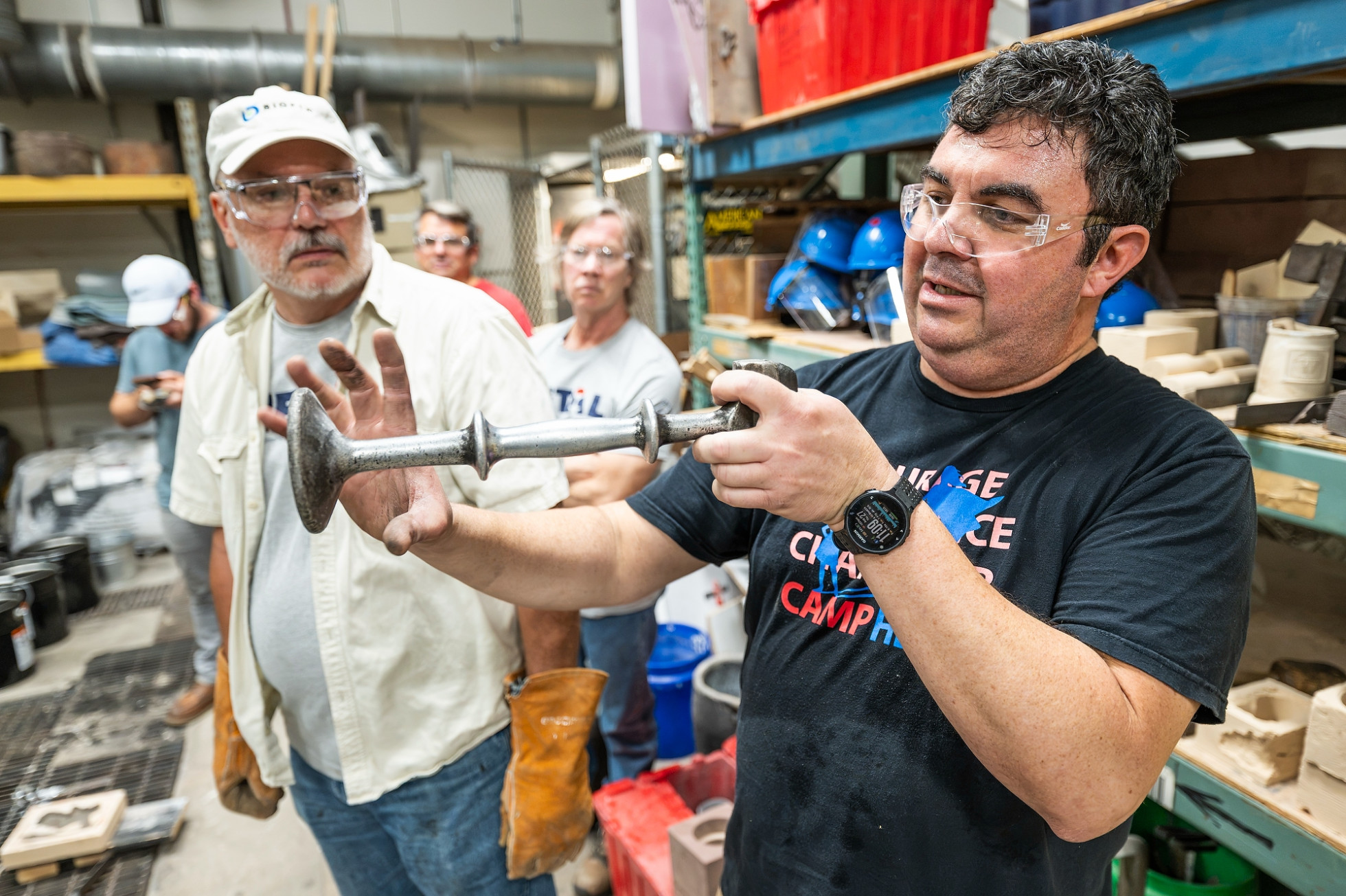 Herrera wears safety glasses and holds a molded piece of aluminum while talking bootcamp participants.