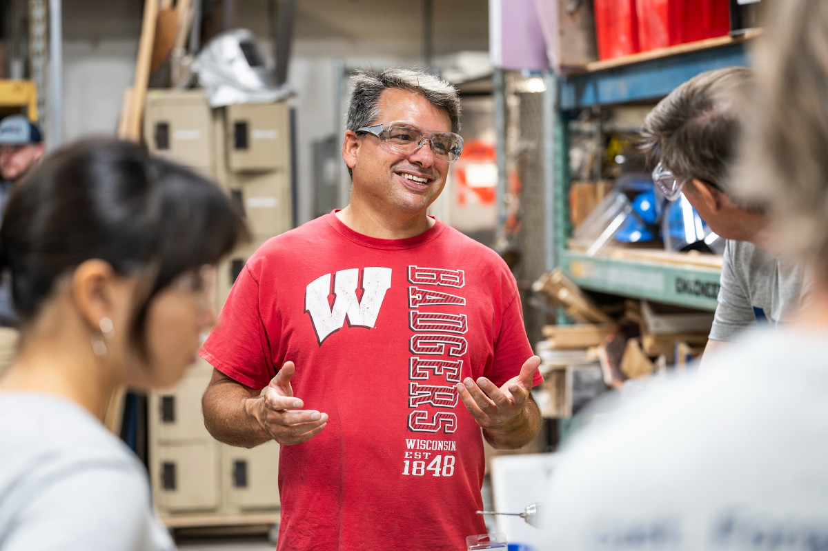 De Cicco wears a red Wisconsin Badgers shirt and safety glasses while talking to a group of METAL bootcamp participants.